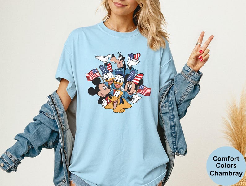 May include: Light blue t-shirt featuring a graphic of Mickey Mouse, Minnie Mouse, Donald Duck, Daisy Duck, Goofy, and Pluto, each holding an American flag. The shirt is a comfort colors chambray.