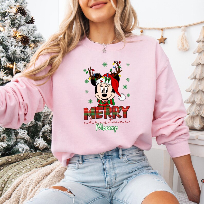 May include: A light pink sweatshirt featuring a Christmas-themed Minnie Mouse graphic. Minnie is wearing a Santa hat and reindeer antlers with Christmas lights. The text "MERRY Christmas Mommp" is printed below the graphic.