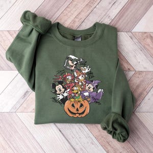 May include: An olive green sweatshirt featuring a Halloween-themed graphic of cartoon characters, including Mickey Mouse, Minnie Mouse, Donald Duck, and Goofy, surrounding a jack-o'-lantern. The characters are in costume.
