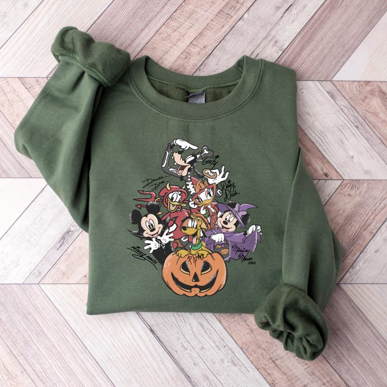 May include: An olive green sweatshirt featuring a Halloween-themed graphic of cartoon characters, including Mickey Mouse, Minnie Mouse, Donald Duck, and Goofy, surrounding a jack-o'-lantern. The characters are in costume.