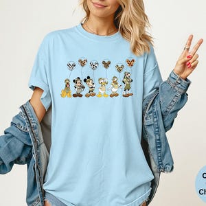 K&ouml;nnte beinhalten: Hellblaues T-Shirt mit den Zeichentrickfiguren Pluto, Mickey Mouse, Donald Duck und Goofy, die jeweils einen gemusterten Ballon halten. Die Ballons haben Zebra-, Leoparden- und Dalmatiner-Prints. Auf dem Shirt steht "Comfort Colors Chambray".