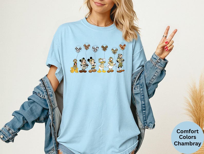 K&ouml;nnte beinhalten: Hellblaues T-Shirt mit den Zeichentrickfiguren Pluto, Mickey Mouse, Donald Duck und Goofy, die jeweils einen gemusterten Ballon halten. Die Ballons haben Zebra-, Leoparden- und Dalmatiner-Prints. Auf dem Shirt steht "Comfort Colors Chambray".