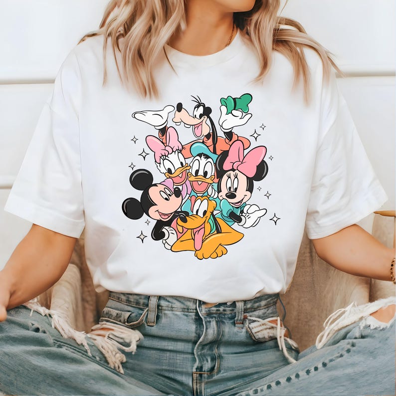 May include: White t-shirt with a colorful graphic print of Mickey Mouse, Minnie Mouse, Donald Duck, Goofy, and Pluto. The characters are arranged in a playful cluster, with Mickey Mouse and Minnie Mouse in the center.