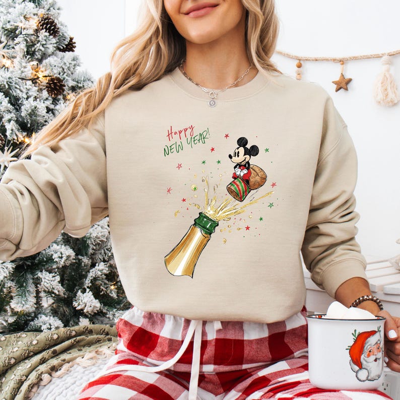 May include: Beige sweatshirt with a festive graphic of Mickey Mouse popping a champagne bottle, with the text "Happy New Year!". The design includes stars and a Santa hat. The sweatshirt is paired with red and white plaid pajama bottoms.