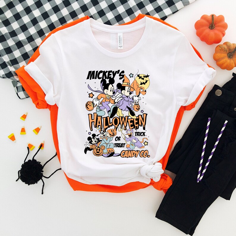 May include: White t-shirt with a graphic of Mickey Mouse and friends dressed in Halloween costumes. The text on the shirt reads "Mickey's Halloween or Treat Candy Co."