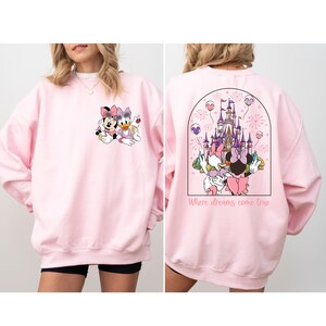 Pu&ograve; includere: Felpa rosa con un'immagine di Topolino e Minnie Mouse sul davanti e un'immagine del castello di Cenerentola con Topolino e Minnie Mouse sul retro. Il testo "Where dreams come true" &egrave; stampato sotto l'immagine del castello.