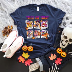 May include: Navy blue t-shirt with a Halloween-themed graphic. The design features the text "Trick or Treat" and images of Disney characters in polaroid-style frames. White sneakers, pumpkins, and a skull are also visible.