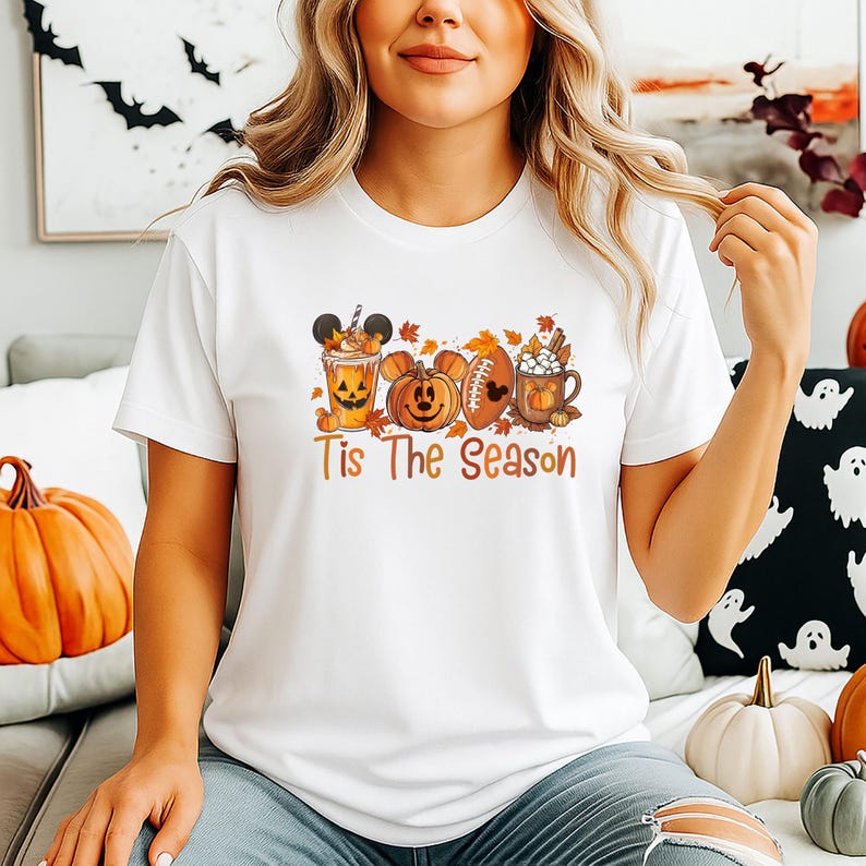 K&ouml;nnte beinhalten: Wei&szlig;es T-Shirt mit einem herbstlichen Grafikdesign. Das Design zeigt einen K&uuml;rbis, einen Football und eine Tasse hei&szlig;e Schokolade, alle mit cartoonartigen Merkmalen. Der Text "Tis The Season" ist in einem warmen Orange gedruckt.