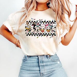 May include: A white t-shirt with a black and white checkered pattern and a row of cartoon characters wearing Santa hats. The characters include Mickey Mouse, Minnie Mouse, Donald Duck, Daisy Duck, and Goofy. The characters are surrounded by a string of Christmas lights.
