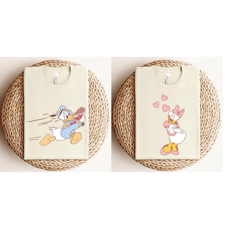 May include: Two beige t-shirts featuring cartoon images of Donald Duck and Daisy Duck. Donald Duck is wearing a sailor hat and holding a baseball bat. Daisy Duck is wearing a pink bow and blowing kisses.
