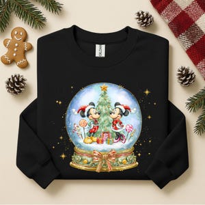 May include: Black sweatshirt with a snow globe design featuring Mickey and Minnie Mouse in festive Christmas outfits. The snow globe includes a Christmas tree, gifts, and candy canes. Gold stars and a bow decorate the base.
