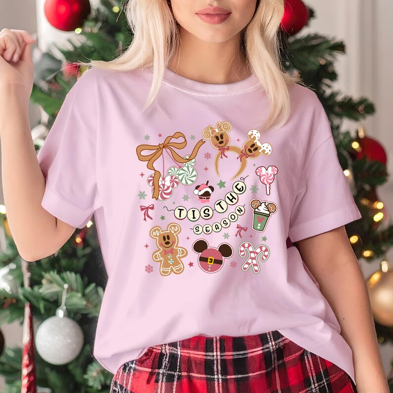 May include: A pale pink t-shirt with a festive graphic featuring gingerbread men, candy canes, and the phrase "Tis the Season." The design includes Mickey Mouse-themed elements and a bow. The shirt is paired with red plaid pajama bottoms.