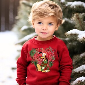 May include: A red sweatshirt with a Christmas-themed design featuring a cartoon dog wearing a green and red sweater. The dog is surrounded by red and green candy canes, snowflakes, and stars.