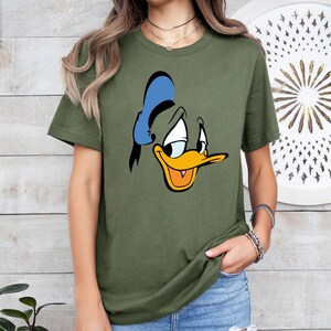 May include: Olive green t-shirt featuring a cartoon image of Donald Duck's face. The graphic includes a blue hat, yellow beak, and a sad expression. The shirt is a casual style.