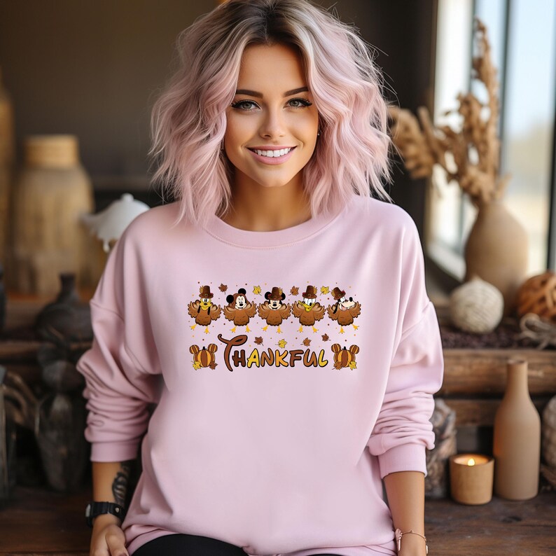May include: A light pink sweatshirt featuring a Thanksgiving-themed design. The design includes cartoon turkeys wearing hats and the word "THANKFUL" in a decorative font. The sweatshirt is a crew neck style.