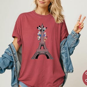 May include: Crimson-colored t-shirt featuring a graphic of the Eiffel Tower with Disney characters. The design includes Mickey and Minnie Mouse, Donald Duck, and Goofy. The shirt is a Comfort Colors brand.