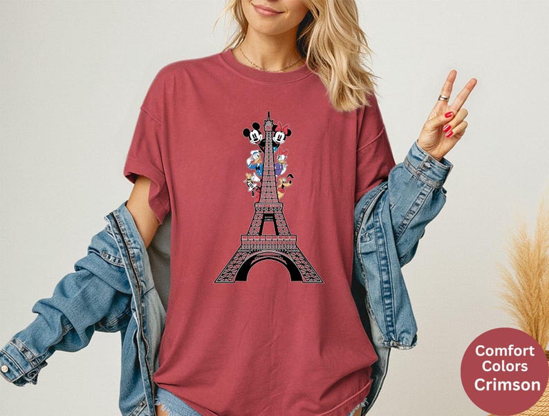 May include: Crimson-colored t-shirt featuring a graphic of the Eiffel Tower with Disney characters. The design includes Mickey and Minnie Mouse, Donald Duck, and Goofy. The shirt is a Comfort Colors brand.