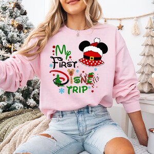 May include: Pink sweatshirt with a Christmas-themed Mickey Mouse design. The text on the sweatshirt reads "My First Disney Trip".