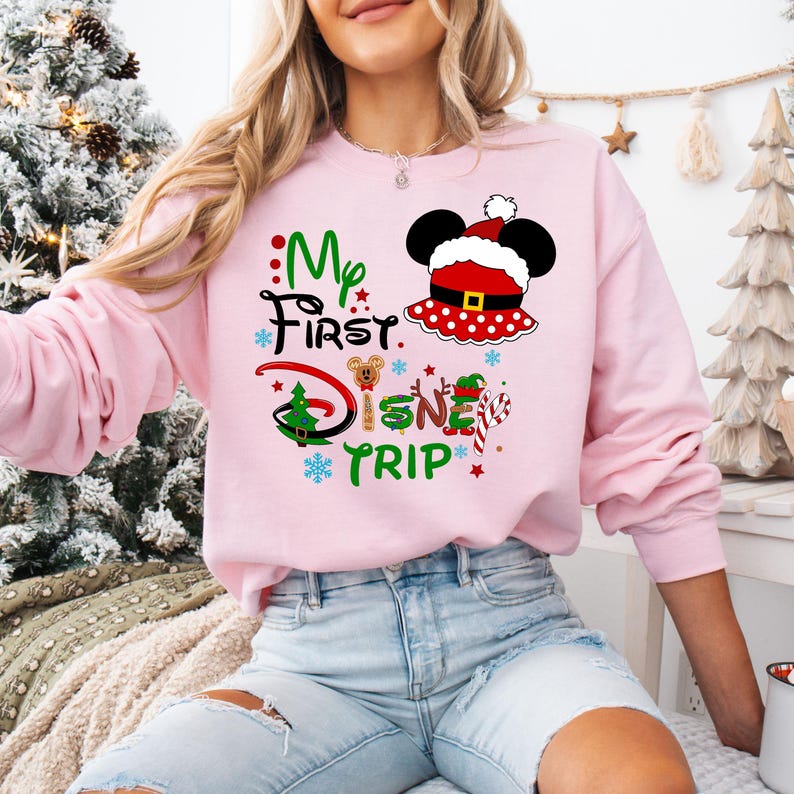 May include: Pink sweatshirt with a Christmas-themed Mickey Mouse design. The text on the sweatshirt reads "My First Disney Trip".