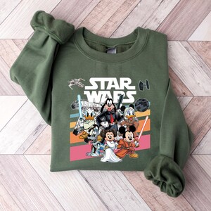 K&ouml;nnte beinhalten: Gr&uuml;nes Sweatshirt mit einem Star Wars-Motiv mit Disney-Figuren. Das Design umfasst Micky Maus, Minnie Maus, Donald Duck, Goofy und andere Charaktere mit Lichtschwertern und Blastern. Die Worte "STAR WARS" sind in wei&szlig;en Blockbuchstaben.