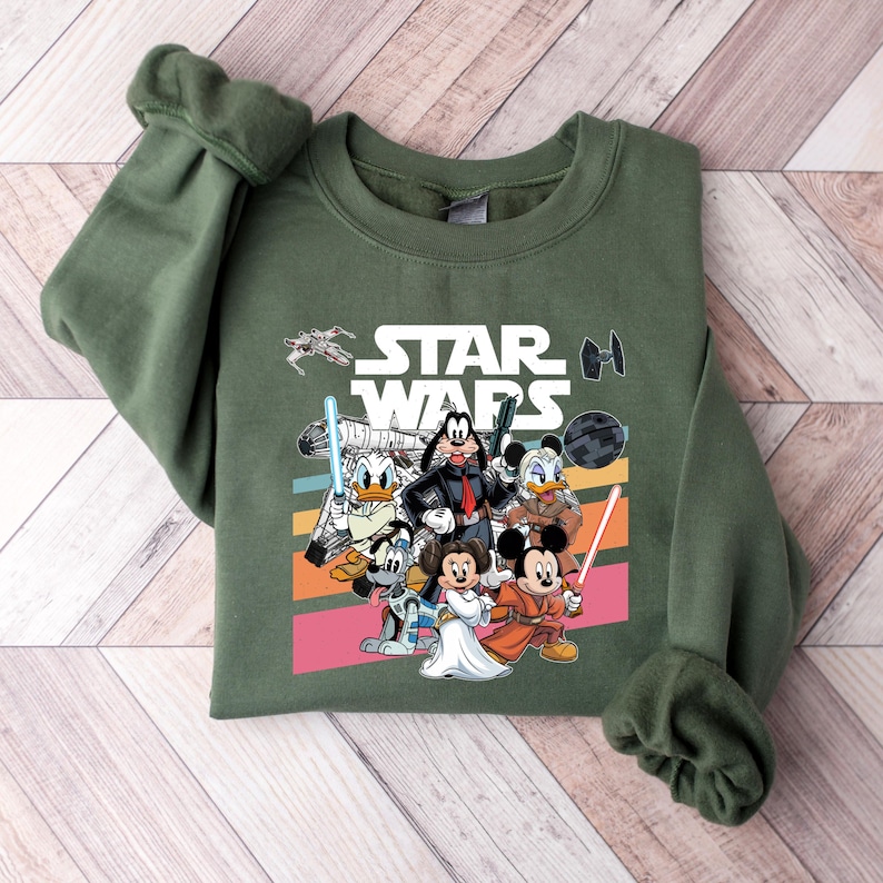 K&ouml;nnte beinhalten: Gr&uuml;nes Sweatshirt mit einem Star Wars-Motiv mit Disney-Figuren. Das Design umfasst Micky Maus, Minnie Maus, Donald Duck, Goofy und andere Charaktere mit Lichtschwertern und Blastern. Die Worte "STAR WARS" sind in wei&szlig;en Blockbuchstaben.