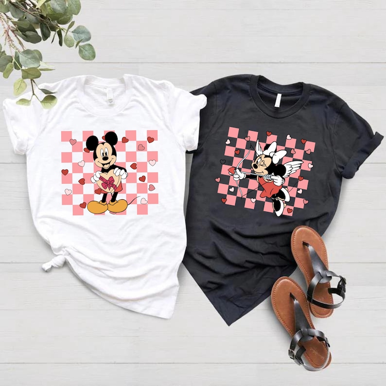 May include: Two t-shirts with a checkered pattern and pink hearts. One shirt features Mickey Mouse holding a heart, the other features Minnie Mouse as Cupid with wings and an arrow.