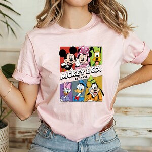 Pu&ograve; includere: Maglietta rosa con una grafica colorata di Topolino e i suoi amici. La grafica include Topolino, Minnie Mouse, Paperino, Pippo, Pluto e Paperina. Il testo "Mickey & Co. EST. 1928" &egrave; stampato sotto la grafica.