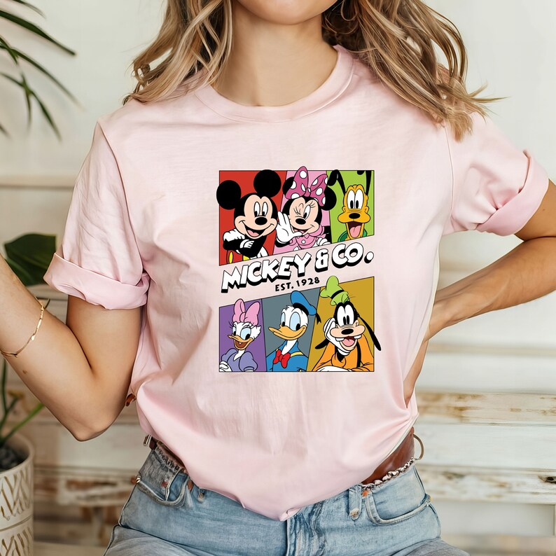 Pu&ograve; includere: Maglietta rosa con una grafica colorata di Topolino e i suoi amici. La grafica include Topolino, Minnie Mouse, Paperino, Pippo, Pluto e Paperina. Il testo "Mickey & Co. EST. 1928" &egrave; stampato sotto la grafica.