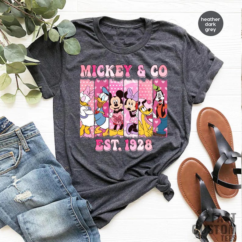 Puede incluir: Una camiseta gris oscuro jaspeada con un gr&aacute;fico rosa y blanco de Mickey Mouse y sus amigos. El gr&aacute;fico incluye el texto "Mickey & Co. Est. 1928".