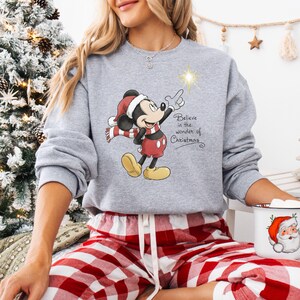 May include: A gray sweatshirt featuring a Christmas-themed Mickey Mouse design. Mickey is wearing a Santa hat and scarf, pointing at a star. The text "Believe in the wonder of Christmas" is also on the sweatshirt.