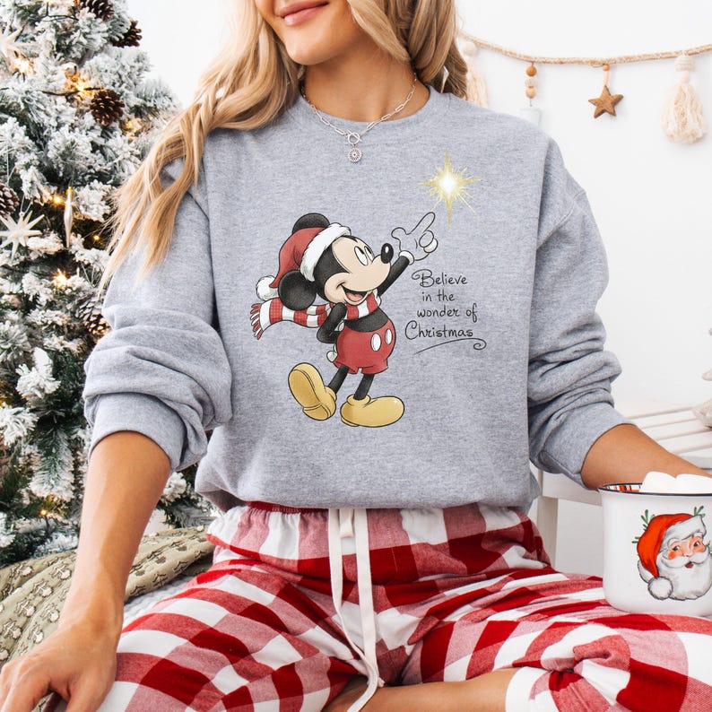 May include: A gray sweatshirt featuring a Christmas-themed Mickey Mouse design. Mickey is wearing a Santa hat and scarf, pointing at a star. The text "Believe in the wonder of Christmas" is also on the sweatshirt.