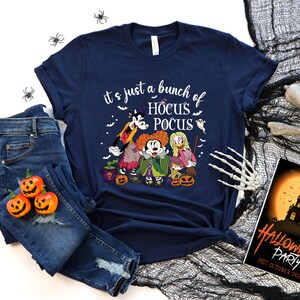 Op de afbeelding: Een marineblauw t-shirt met een afbeelding van Mickey Mouse, Donald Duck en Winnie de Poeh verkleed als heksen. De tekst "it's just a bunch of HOCUS POCUS" staat op het shirt gedrukt.