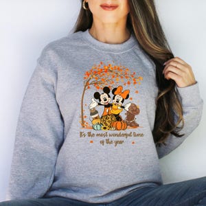May include: A grey sweatshirt with a graphic of Mickey and Minnie Mouse in autumn outfits, positioned near pumpkins and a tree with orange leaves. The text "It's the most wonderful time of the year" is printed below the graphic.
