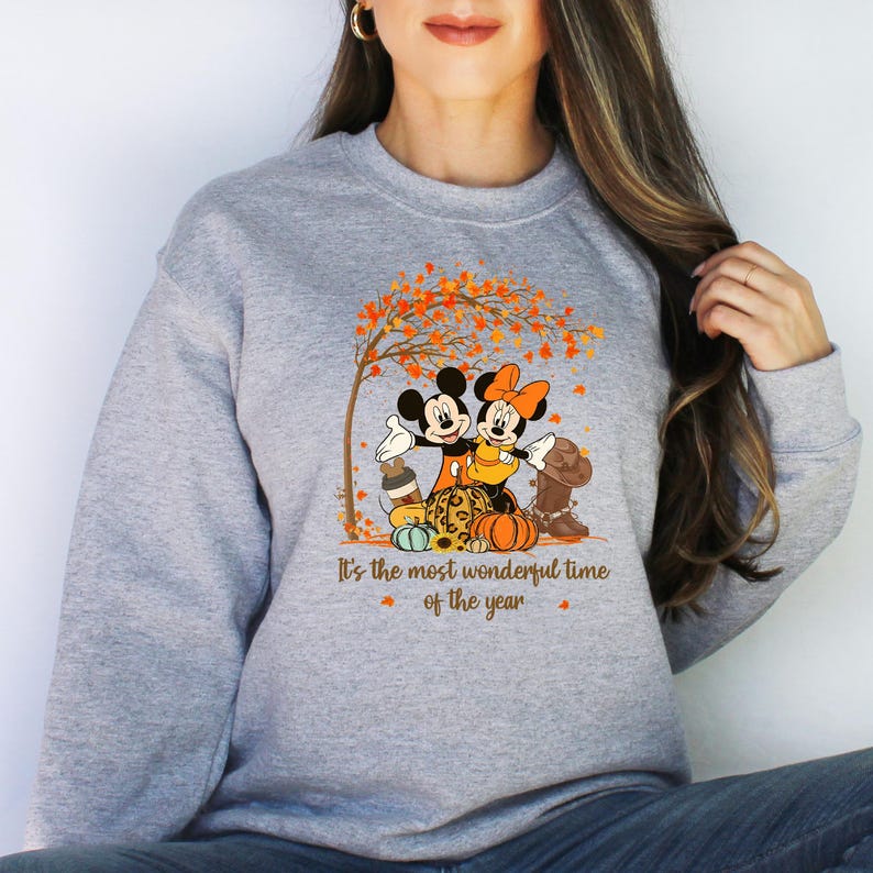 May include: A grey sweatshirt with a graphic of Mickey and Minnie Mouse in autumn outfits, positioned near pumpkins and a tree with orange leaves. The text "It's the most wonderful time of the year" is printed below the graphic.