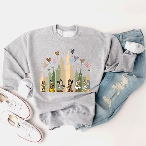 May include: A gray sweatshirt with a graphic print of Mickey Mouse, Donald Duck, Goofy, and Pluto in front of a castle silhouette. The characters are wearing safari outfits and there are colorful balloons in the air.
