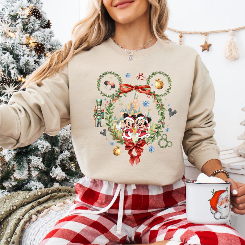 May include: Beige sweatshirt featuring a festive Mickey and Minnie Mouse design. The graphic includes a wreath, castle, and snowflakes. The sweatshirt is paired with red and white plaid pajama pants. A white mug with a Santa Claus graphic is also visible.