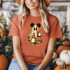 May include: Orange t-shirt featuring a cartoon ghost character with Mickey Mouse ears, holding a jack-o'-lantern. The ghost is dressed in a patchwork quilt-like costume. The shirt is a solid orange color, perfect for Halloween.