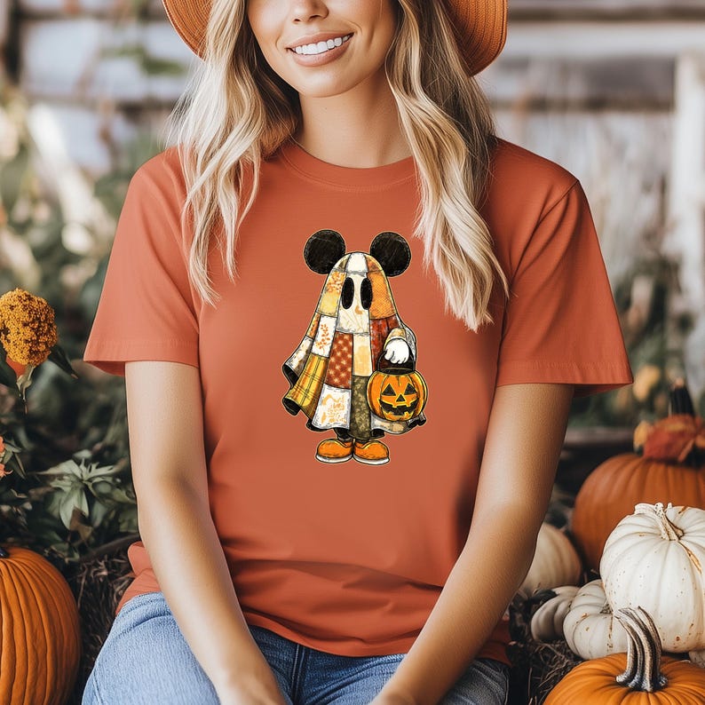 May include: Orange t-shirt featuring a cartoon ghost character with Mickey Mouse ears, holding a jack-o'-lantern. The ghost is dressed in a patchwork quilt-like costume. The shirt is a solid orange color, perfect for Halloween.