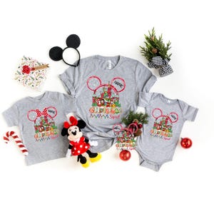 Puede incluir: Camisetas grises de "Christmas Squad" y un body con un dise&ntilde;o de Mickey Mouse. Las camisetas presentan un gr&aacute;fico de castillo, luces navide&ntilde;as y el texto "Christmas Squad". Tambi&eacute;n hay un peluche de Minnie Mouse y orejas de Mickey Mouse.