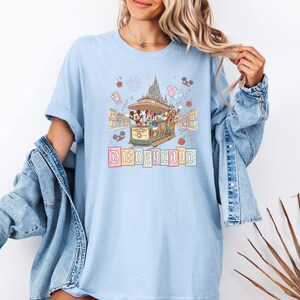 May include: Light blue t-shirt featuring a Disneyland graphic. The design includes a trolley with cartoon characters, a castle, and the word "Disneyland" in pastel colors. The shirt has short sleeves and a relaxed fit.