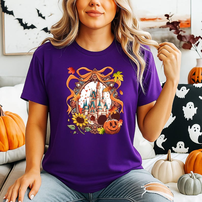 May include: A purple t-shirt featuring a colorful graphic of a castle, sunflowers, and a jack-o'-lantern. The graphic is framed with a decorative ribbon and autumn leaves. The shirt is a short-sleeved crew neck.