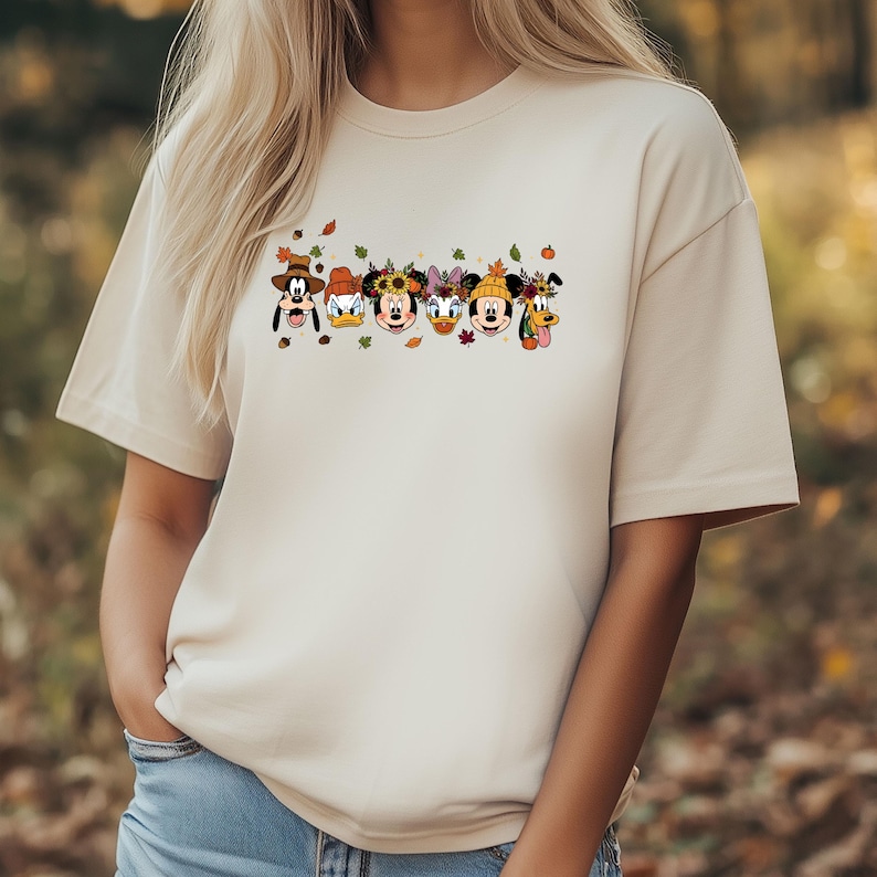 May include: Beige t-shirt featuring a graphic of Disney characters, including Goofy, Donald Duck, Minnie Mouse, Daisy Duck, Mickey Mouse, and Pluto, each adorned with fall-themed accessories. The design includes autumn leaves.