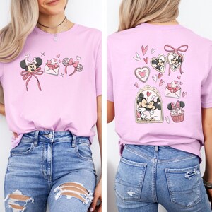 May include: A light pink t-shirt featuring Minnie Mouse-themed graphics. The front displays Minnie with a bow, hearts, and a headband. The back showcases Minnie in a heart-shaped frame, a cupcake, and other heart-themed designs. The shirt is made of a soft material.