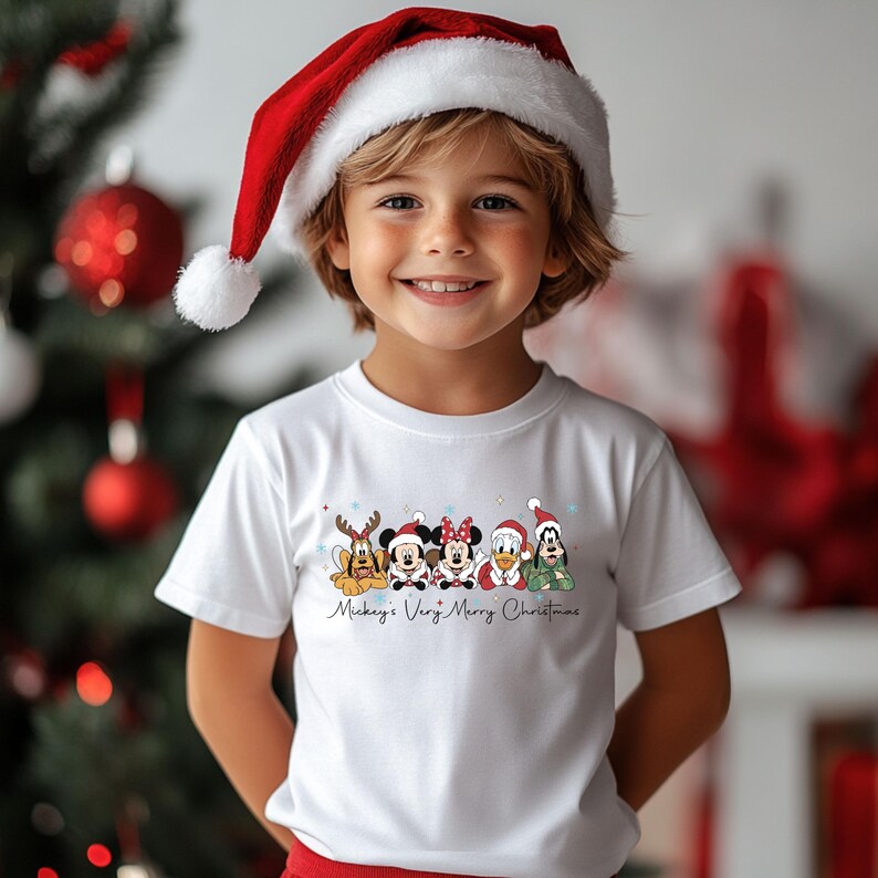 Puede incluir: Un ni&ntilde;o lleva una camiseta blanca con personajes de Disney vestidos con atuendos navide&ntilde;os. La camiseta tiene el texto "Mickey's Very Merry Christmas". El ni&ntilde;o tambi&eacute;n lleva un gorro de Pap&aacute; Noel rojo con un pomp&oacute;n blanco.