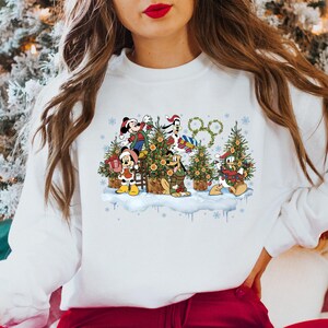 May include: White sweatshirt with a Christmas-themed design featuring Mickey Mouse, Donald Duck, Goofy, and Pluto decorating a Christmas tree. The design includes a wreath with the iconic Disney logo.