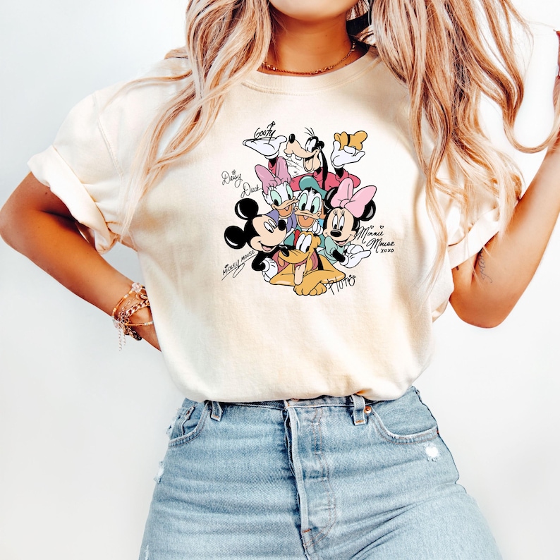 May include: Cream-colored t-shirt featuring a colorful graphic of Disney characters including Mickey Mouse, Minnie Mouse, Donald Duck, Daisy Duck, Goofy, and Pluto. The graphic includes character signatures and the text "Minnie Mouse 2020".