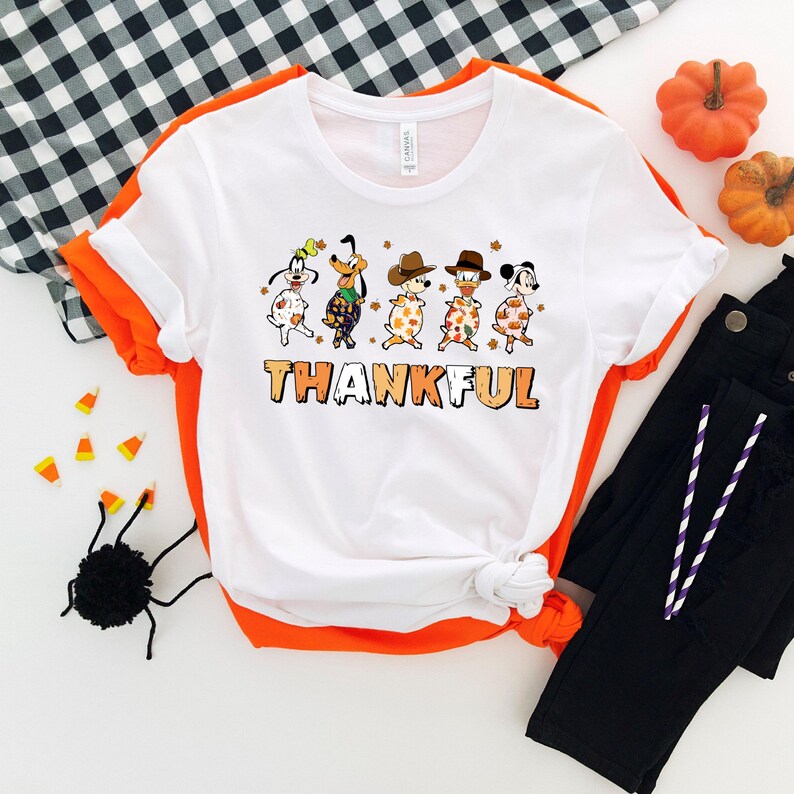 May include: White t-shirt with orange trim featuring cartoon dogs in fall attire. The word "THANKFUL" is printed in orange and brown. The shirt is styled with black pants, pumpkins, and Halloween decorations.