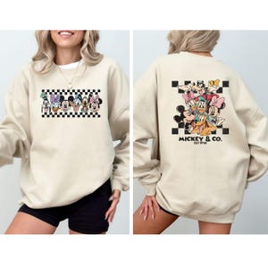May include: Beige sweatshirt featuring a cartoon graphic of Mickey Mouse and friends on the front and back. The front graphic includes a checkered border. The back graphic has the text "Mickey & Co. Est 1928".