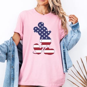May include: A pale pink t-shirt with a Mickey Mouse silhouette filled with the American flag design. The shirt has short sleeves and a loose fit, suitable for everyday wear. The design incorporates the stars and stripes of the United States flag.