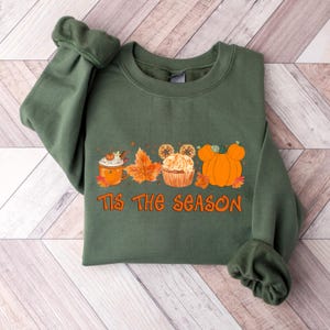 Pu&ograve; includere: Felpa verde con grafica arancione con la scritta "Tis The Season". La grafica include un latte speziato alla zucca, foglie d'autunno, un cupcake con orecchie di Topolino e zucche. Un capo a tema autunnale.